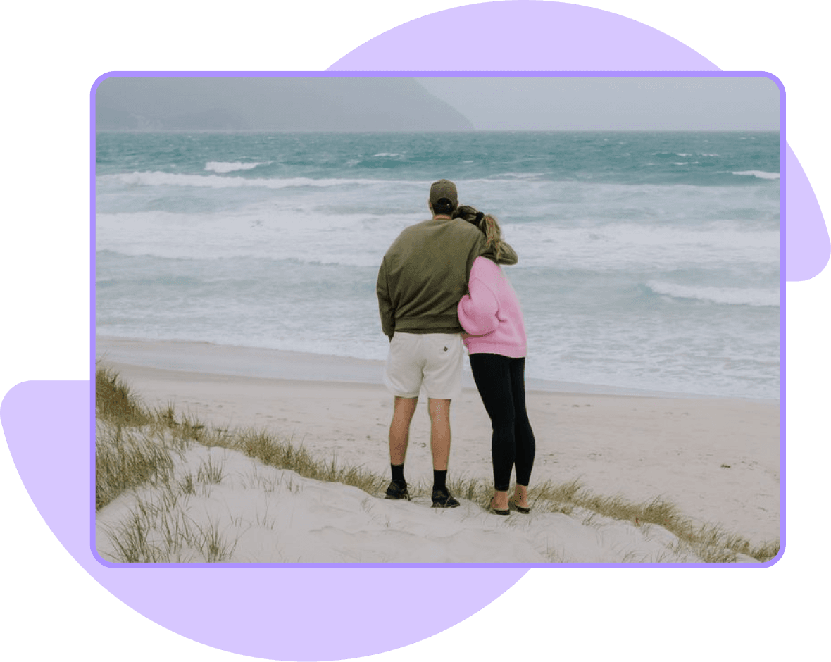 Two people embracing while looking at the ocean from a sandy beach.