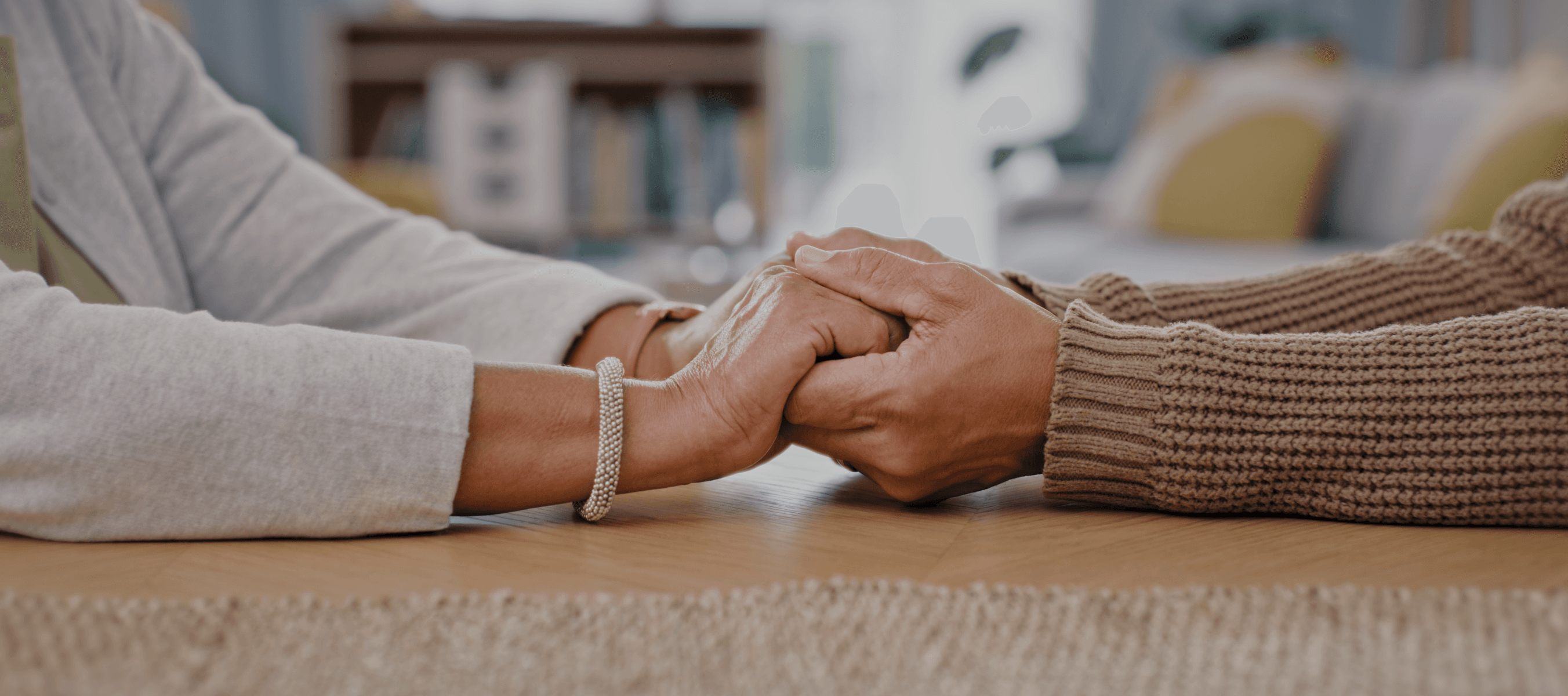 Two people holding hands on a wooden table, expressing comfort and solidarity.