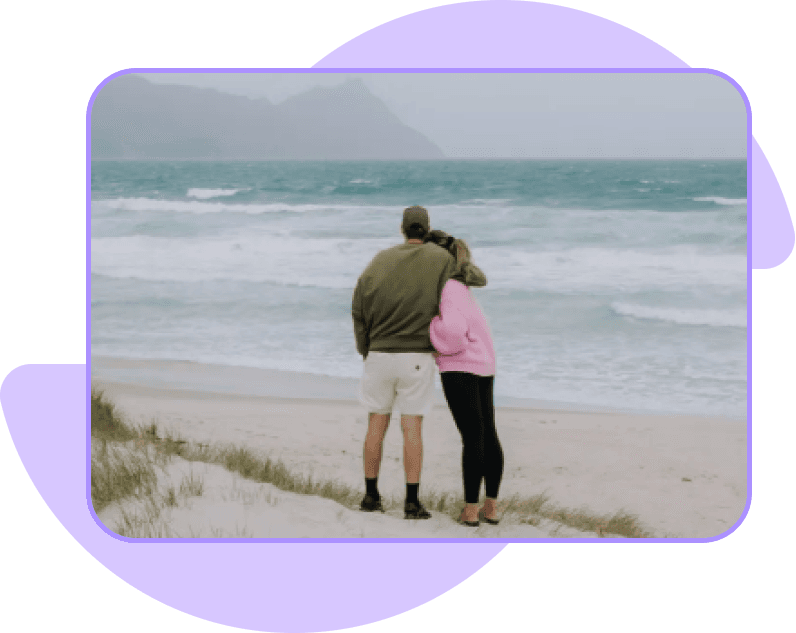 Two people embracing while looking at the ocean from a sandy beach.